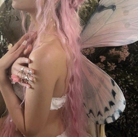 a white feminine person with long pink hair and butterfly-like fairy wings. their face is off camera and they are hudding themselves with their arms. they are weraing multiple silver rings and a white bandou top