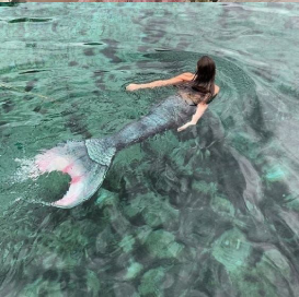 a white feminine person with long brown hair and teal mermaid tail is swimming in a clear blue body of water.