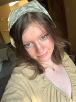 a selfie of melanie, a white feminine person with dark blue eyes and blackish-brown hair with blonde streaks. they are wearing a sligthly sheer lacy white bandana, a golden necklace with a chai charm and a light tan dress with sleves and a v-neck.